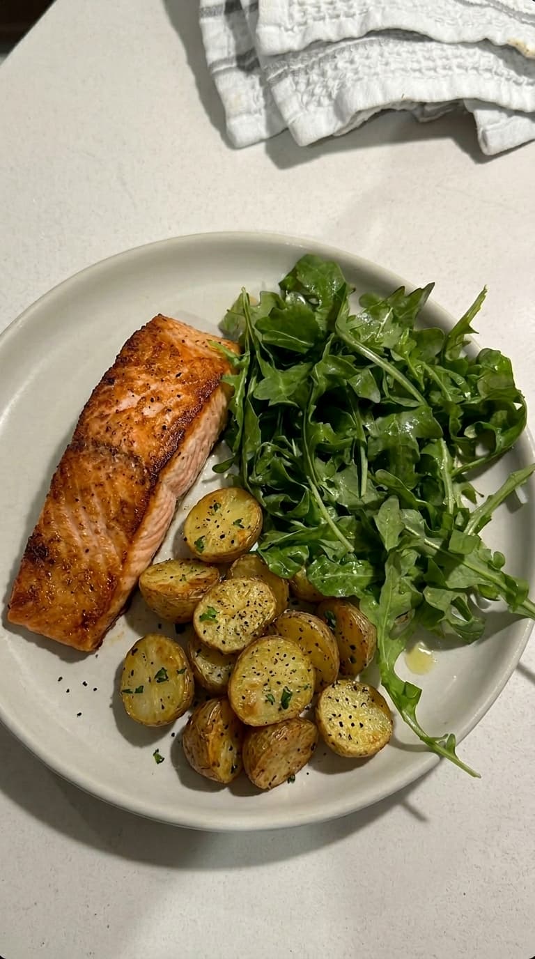 A plate with a piece of grilled salmon, roasted potato slices, and a side of fresh arugula salad.