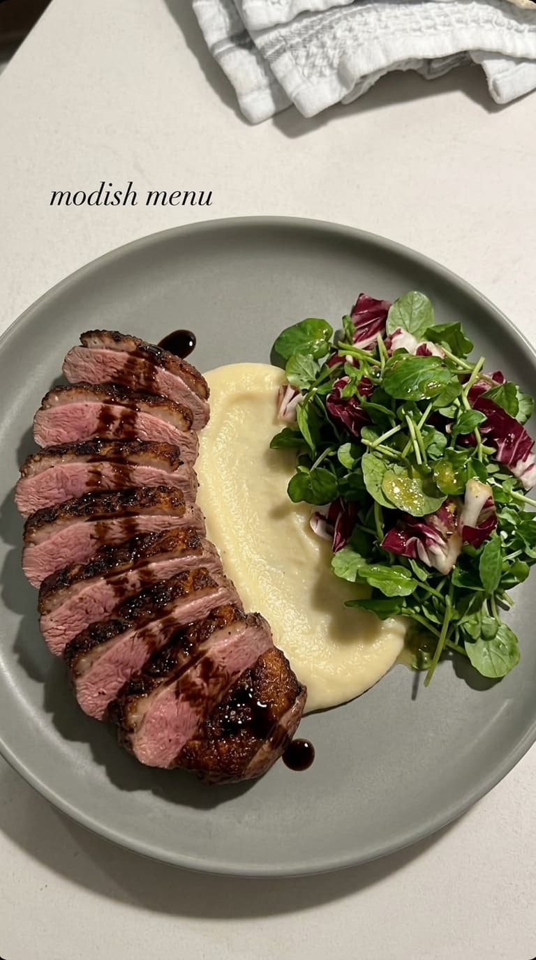 Sliced seared duck breast with pink center, drizzled with sauce, served with apple puree and a green and red salad on a grey plate.